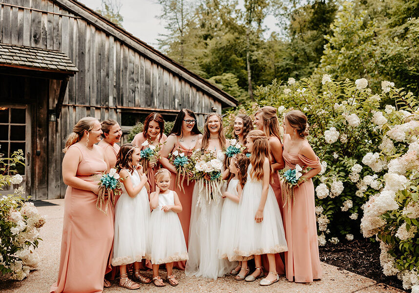 2025_DWC_Weddings_BridesMaids_1 Bride's Maids in soft colors standing in front of the Dogwood Canyon Mill with the flower girls.